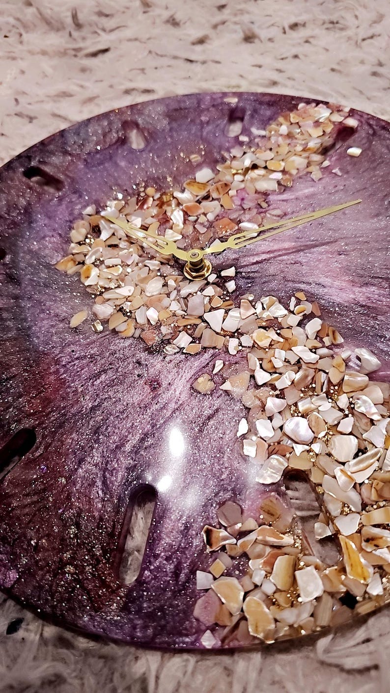 May include: A circular wall clock featuring a deep purple and metallic swirl resin base. An S-shaped band of crushed iridescent seashells and gold glitter runs across the center. The clock has thin gold-toned metal hour and minute hands.