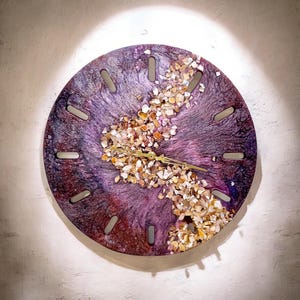 May include: A round wall clock with a purple and maroon resin background. The clock face features a stream of small, light-colored shells and gold-colored clock hands. The clock has 12 rectangular markers around the edge.