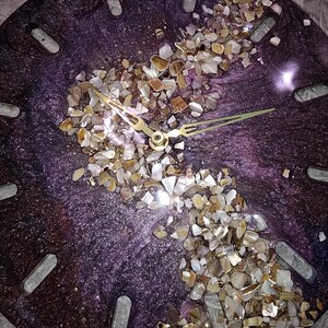 May include: A round wall clock with a deep purple, shimmering resin base featuring a diagonal band of crushed, multi-colored stone and shell fragments. The clock face includes gold-toned metal hands and rectangular cutouts for hour markers.