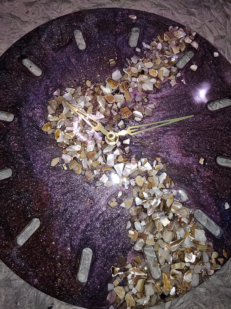 May include: A round wall clock with a deep purple, shimmering resin base featuring a diagonal band of crushed, multi-colored stone and shell fragments. The clock face includes gold-toned metal hands and rectangular cutouts for hour markers.