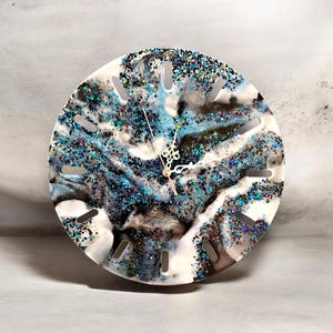 May include: Round wall clock with a unique design. The clock face features a swirling pattern of white, blue, and black resin, accented with glitter. The clock has a silver-tone clock hands and hour markers. The clock measures approximately 10 inches in diameter.