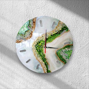 May include: A circular wall clock featuring a white resin base with a textured, geode-inspired design. It includes vibrant green and gold accents, embedded glass crystals, and thin black clock hands with a red second hand on a white face.