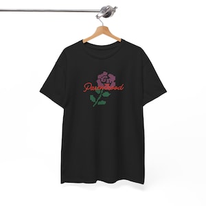 May include: Black t-shirt with a floral design and the word "Parenthood" in red script. The flower is made of pink and purple pixels, with green pixelated leaves and stem. The shirt is hanging on a wooden hanger.