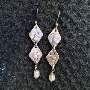 May include: A pair of handcrafted earrings with a diamond-shaped design. Each earring has two diamond-shaped pendants with etched patterns and a small pearl at the bottom. The earrings are made of a copper-coloured metal.
