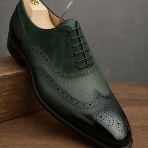 May include: A dark green leather dress shoe with a wingtip design. The shoe features a textured leather upper, a suede panel, and a glossy toe cap. Black laces and a wooden shoe tree are visible.