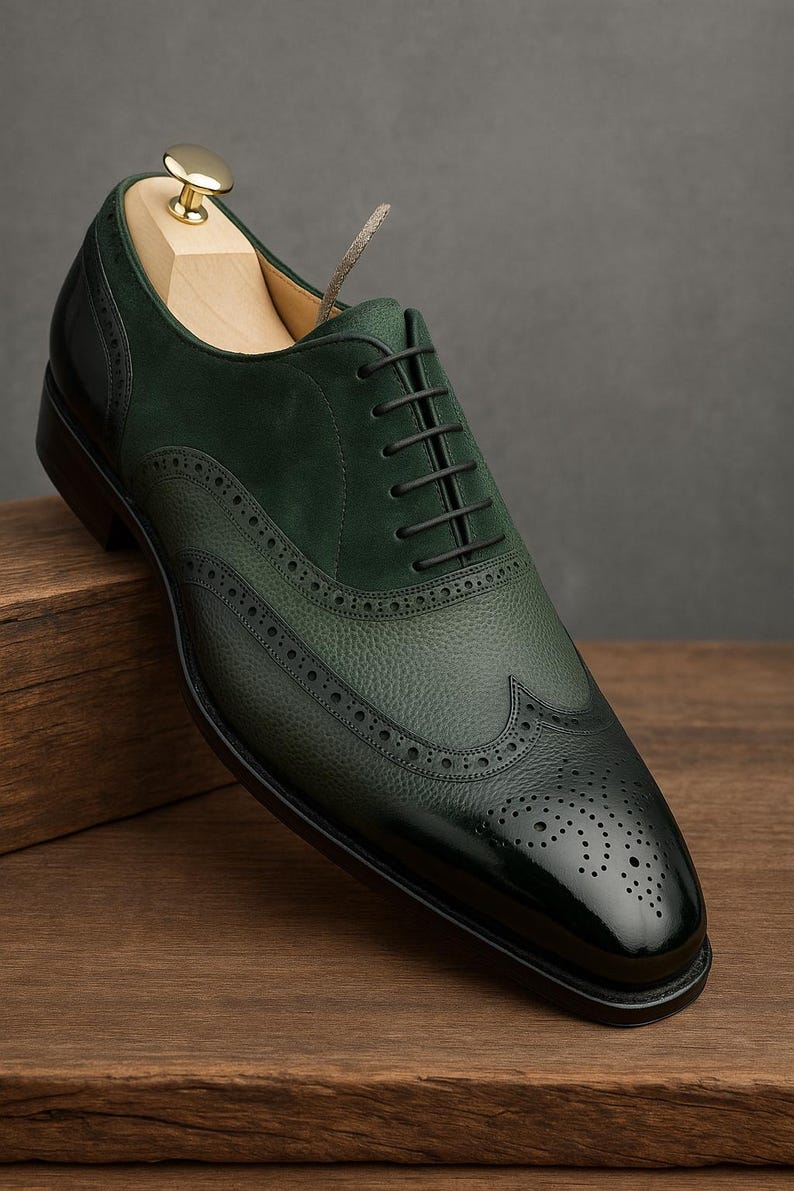 May include: A dark green leather dress shoe with a wingtip design. The shoe features a textured leather upper, a suede panel, and a glossy toe cap. Black laces and a wooden shoe tree are visible.