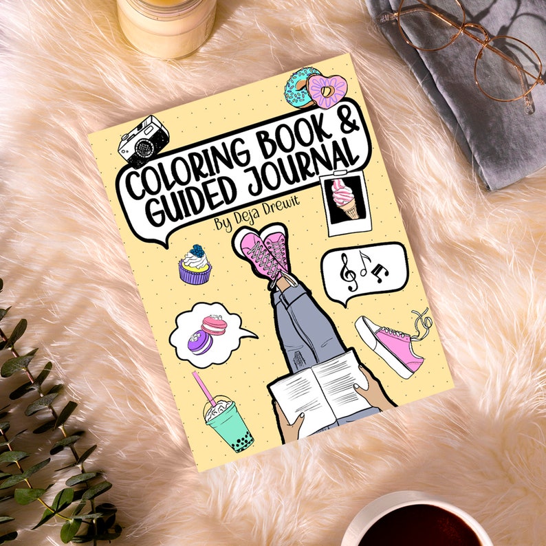 May include: A yellow colouring book and guided journal cover with a cartoon illustration of a person reading a book with their legs in the air. The cover also features a camera, ice cream cone, cupcakes, macarons, music notes, and bubble tea.