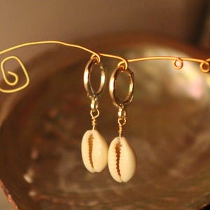 May include: A pair of earrings featuring small, cream-colored cowrie shells suspended from gold-colored hoops. The earrings are displayed against a blurred, neutral background, highlighting the natural texture and shape of the shells.