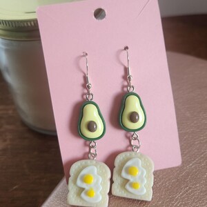May include: A pair of drop earrings featuring a miniature avocado charm with a brown stone, connected by a silver-toned metal link to a charm shaped like a slice of toast topped with two fried eggs, displayed on a rectangular pink card.