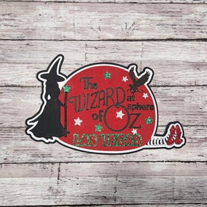 May include: A decorative sticker featuring a black silhouette of a witch with a hat and staff, a red circle with the text "The Wizard of Oz" and "Las Vegas", a black bird, and red glitter shoes.