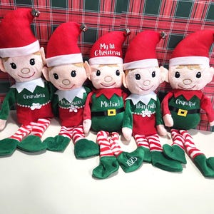 May include: Five plush elf dolls wearing red hats with silver bells, green shirts with white collars, and red and white striped legs. The shirts are embroidered with the names Grandma, Madison, Michael, Hannah, and Grandpa, plus 2025.