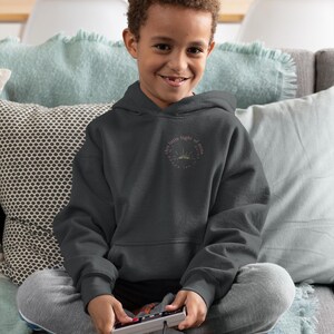 May include: A dark gray hoodie with the text "this little light of mine" in a circular design. The hoodie is worn by a child sitting on a couch, holding a game controller. The child is wearing gray sweatpants and orange socks.