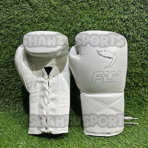 Pu&ograve; includere: Un paio di guantoni da boxe bianchi con la scritta "FLY" e una figura alata stampata sul retro. I guanti sono in pelle e hanno una chiusura con lacci. I guanti sono su una superficie verde.