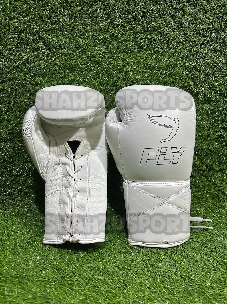 Pu&ograve; includere: Un paio di guantoni da boxe bianchi con la scritta "FLY" e una figura alata stampata sul retro. I guanti sono in pelle e hanno una chiusura con lacci. I guanti sono su una superficie verde.