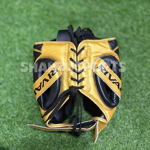 Puede incluir: Un protector de cabeza de boxeo dorado y negro con la palabra "RIVAL" impresa en los laterales. El protector de cabeza tiene cordones y acolchado negros. El texto "SHA SPORTS" es parcialmente visible en el lateral.