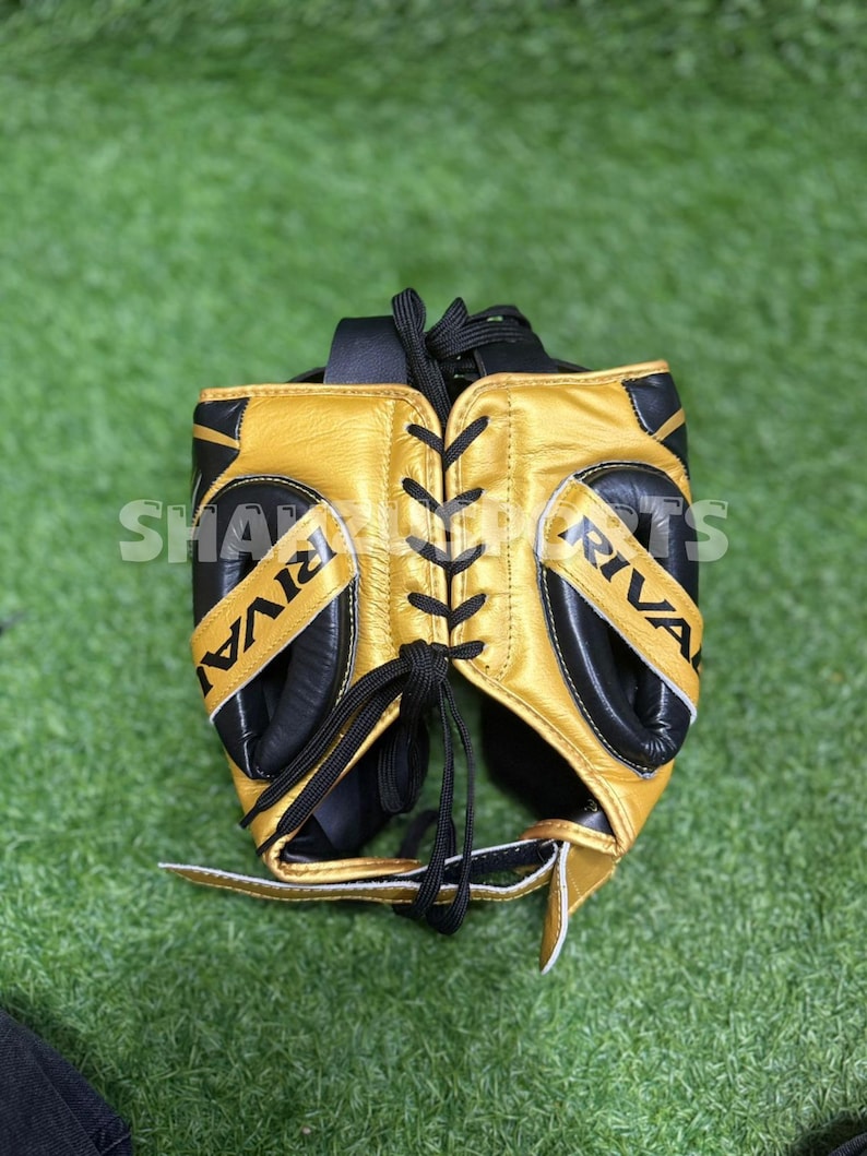 Puede incluir: Un protector de cabeza de boxeo dorado y negro con la palabra "RIVAL" impresa en los laterales. El protector de cabeza tiene cordones y acolchado negros. El texto "SHA SPORTS" es parcialmente visible en el lateral.