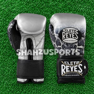 Peut inclure: Une paire de gants de boxe argentés et noirs avec un motif peau de serpent sur la partie supérieure. Les gants arborent le logo Cleto Reyes et l'inscription Hecho en Mexico. Une sangle de poignet noire est présente avec le nom de la marque.