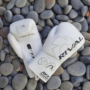 Könnte beinhalten: Ein Paar weiße Boxhandschuhe mit dem Markennamen "RIVAL" in Schwarz aufgedruckt. Die Handschuhe sind aus Leder und tragen den Text "Handcrafted with Pride". Die Handschuhe liegen auf einem Bett aus grauen Steinen.