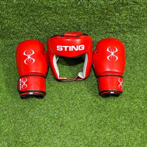 May include: A set of red boxing equipment, including a head guard and two gloves, is displayed on a green surface. The head guard and gloves have white accents and the word "STING" is printed on the head guard.