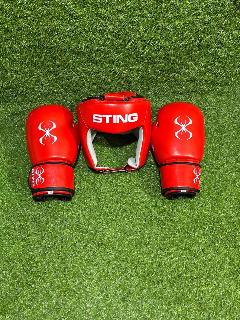 May include: A set of red boxing equipment, including a head guard and two gloves, is displayed on a green surface. The head guard and gloves have white accents and the word "STING" is printed on the head guard.