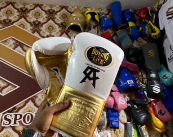 May include: White and gold boxing gloves with the text "NO BOXING NO LIFE" and a logo. The gloves are held up in front of a wall displaying various boxing equipment, including headgear and other gloves.