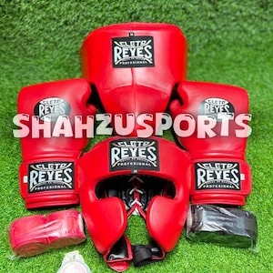 Peut inclure: Un ensemble d'équipement de boxe rouge comprenant deux gants de boxe, un casque avec fermeture à lacets, deux rouleaux de bandes de main et un protège-dents. Chaque article porte le logo Cleto Reyes et le texte SHAHZUSPORTS est superposé.