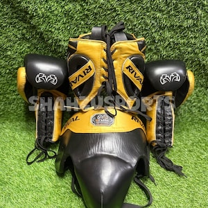 Puede incluir: Un conjunto de boxeo con protector de cabeza, guantes y protector de ingle dorados y negros. La palabra "RIVAL" est&aacute; impresa en el protector de cabeza y en los guantes. El conjunto se muestra sobre una superficie verde.
