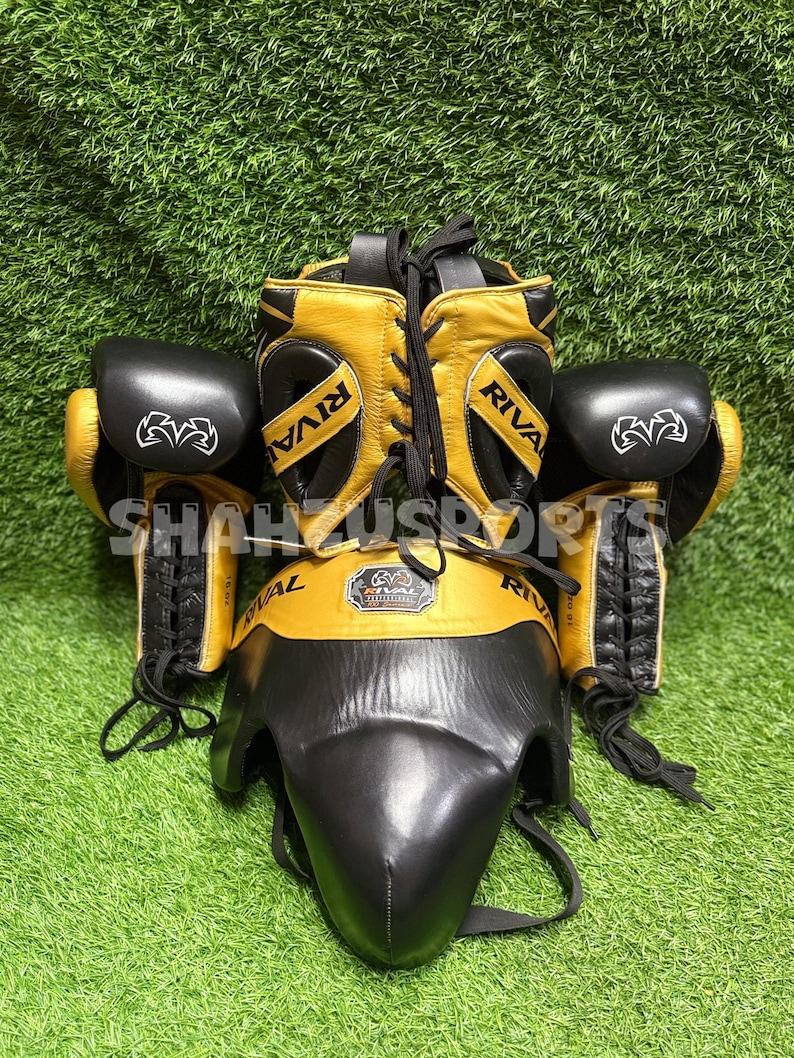 Puede incluir: Un conjunto de boxeo con protector de cabeza, guantes y protector de ingle dorados y negros. La palabra "RIVAL" est&aacute; impresa en el protector de cabeza y en los guantes. El conjunto se muestra sobre una superficie verde.