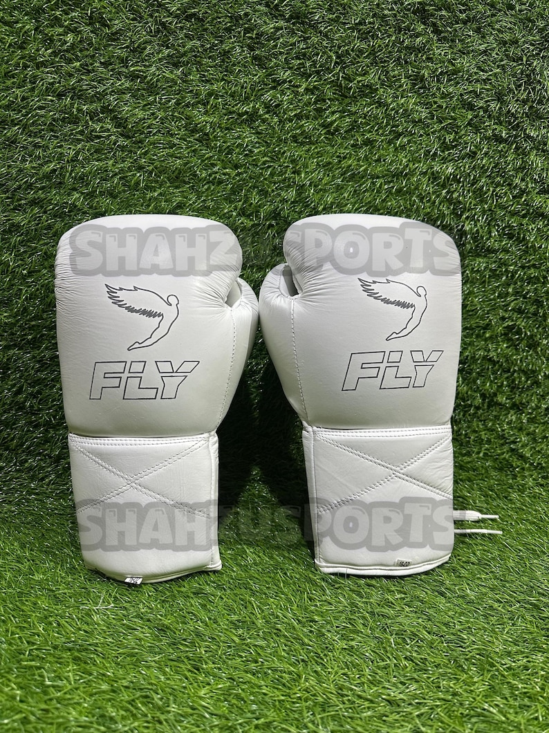 Pu&ograve; includere: Un paio di guantoni da boxe bianchi con la scritta "FLY" stampata in nero. Ogni guanto presenta una grafica di una figura alata. I guantoni sono su uno sfondo di erba verde.