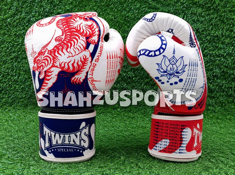 Puede incluir: Un par de guantes de boxeo blancos con gr&aacute;ficos en rojo y azul. El guante izquierdo muestra una ilustraci&oacute;n de un tigre rojo y el texto SHAHZUSPORTS. El guante derecho presenta un dise&ntilde;o de loto azul. Ambos tienen el logo TWINS SPECIAL.