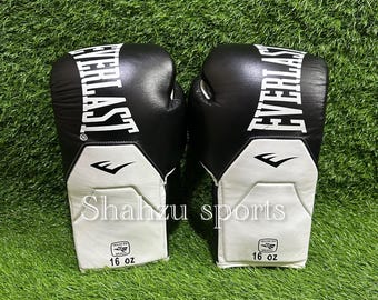 May include: A pair of black and white boxing gloves, each with "EVERLAST" in white. The gloves have a white wrist section with "16 oz" and a logo. The gloves are set against a green background.