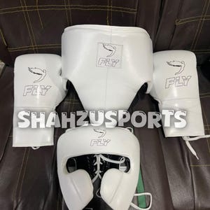 Puede incluir: Un conjunto de equipo de boxeo blanco que incluye dos guantes de boxeo, una coquilla y un casco, todos con el logotipo negro FLY y un gráfico de pájaro. El texto SHAHZUSPORTS está superpuesto en el centro del equipo de cuero blanco.