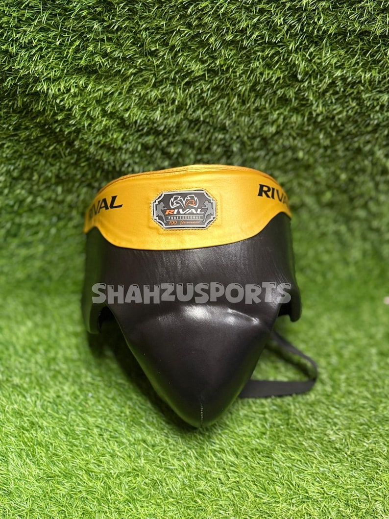 Puede incluir: Protector atl&eacute;tico negro y amarillo con el texto "RIVAL" en la banda amarilla. La copa tiene una carcasa protectora negra y una banda amarilla con el logotipo de la marca. El texto "Shahzusports" est&aacute; impreso en la copa.