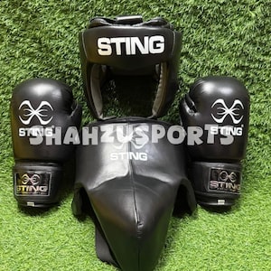 Op de afbeelding: Een zwarte bokshoofdbeschermer met de witte tekst STING, een zwart borstschild met de witte tekst STING en twee zwarte bokshandschoenen met het witte STING-logo en de tekst. De uitrusting heeft een gladde afwerking en witte branding.