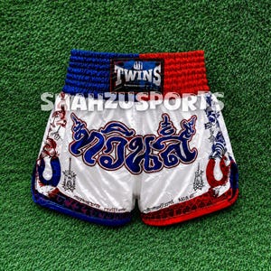 Puede incluir: Pantalones cortos de Muay Thai blancos con cintura azul y roja y un parche negro de Twins. La parte delantera muestra grandes caracteres tailandeses azules, detalles laterales en forma de herradura roja y azul, y gr&aacute;ficos negros. Texto SHAHZUSPORTS visible.