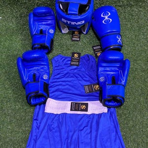 May include: A complete blue boxing set, including a head guard with "STING" text, boxing gloves, a tank top, and boxing shorts with a white waistband. The gloves have a stylized infinity symbol. The set is arranged on a green surface.