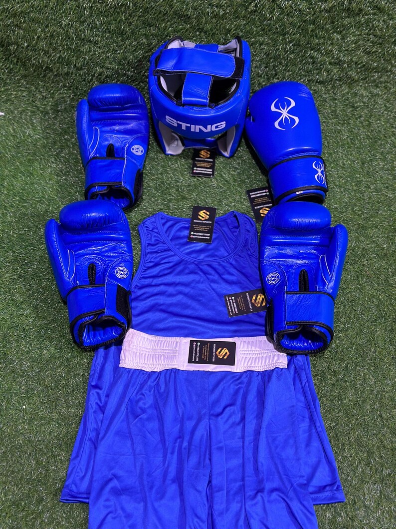 May include: A complete blue boxing set, including a head guard with "STING" text, boxing gloves, a tank top, and boxing shorts with a white waistband. The gloves have a stylized infinity symbol. The set is arranged on a green surface.
