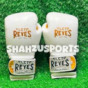 Peut inclure: Une paire de gants de boxe blancs avec une inscription dorée Cleto Reyes Est 1945. Les sangles de poignet présentent une étiquette blanche et dorée avec le texte Cleto Reyes Profesional Hecho en Mexico. Le mot SHAHZUSPORTS est imprimé en blanc.