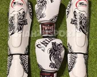 Könnte beinhalten: Weiße Boxhandschuhe und Schienbeinschützer mit schwarzen Drachendesigns. Die Handschuhe und Schienbeinschützer tragen das Logo "Twins Special". Die Handschuhe sind übereinander gestapelt, mit einem rot-schwarzen Band dazwischen. Die Schienbeinschützer befinden sich auf beiden Seiten.