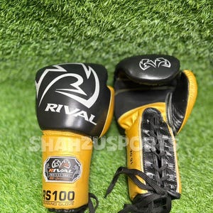Puede incluir: Par de guantes de boxeo Rival RS100 negros y dorados, 16 oz. Los guantes tienen logotipos y texto en blanco y negro. Los guantes est&aacute;n sobre una superficie verde.