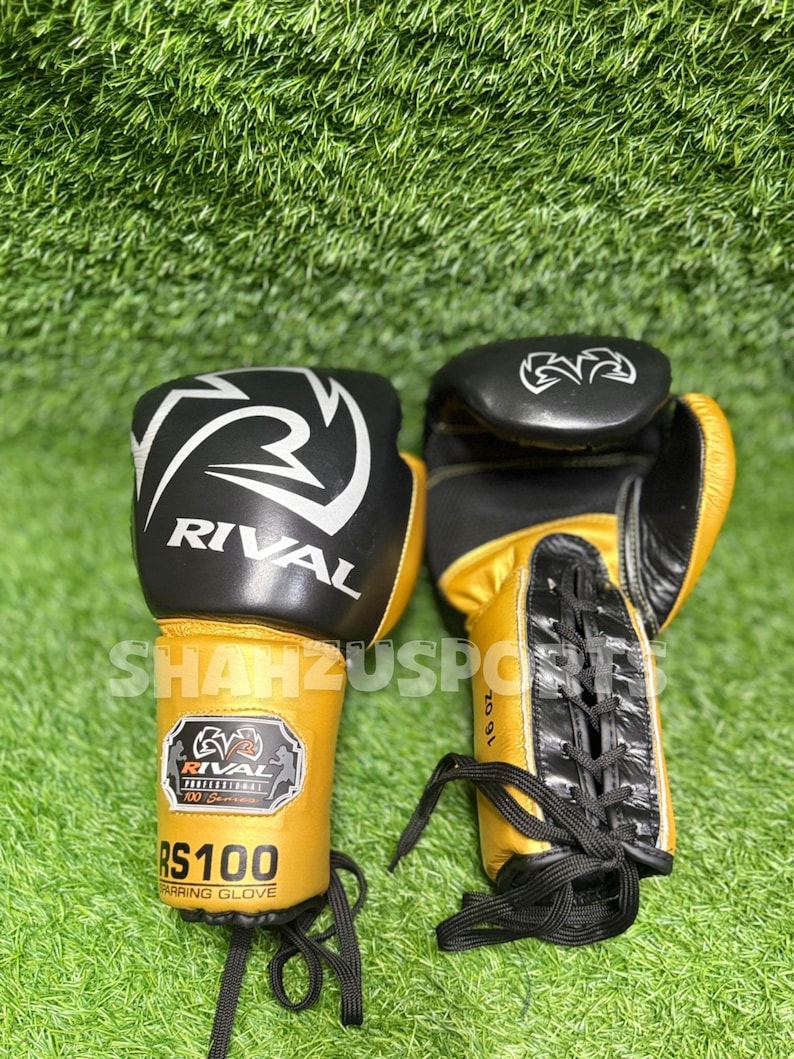 Puede incluir: Par de guantes de boxeo Rival RS100 negros y dorados, 16 oz. Los guantes tienen logotipos y texto en blanco y negro. Los guantes est&aacute;n sobre una superficie verde.