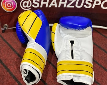 May include: Pair of boxing gloves. The gloves are white, blue, and yellow. The top of the gloves are blue, with yellow and black stripes around the wrist area. The gloves are on a red surface.