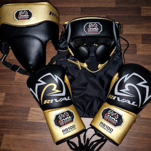 Op de afbeelding: Een set zwarte en gouden boksuitrusting inclusief een hoofdbeschermer, een tok en een paar RS100 sparringhandschoenen. Elk item is voorzien van het Rival Professional 100 Series logo. De handschoenen zijn gelabeld met RS100 Sparring Glove in witte tekst.