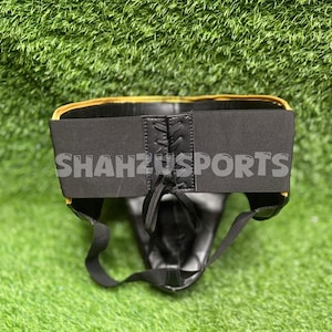 Puede incluir: Protector atl&eacute;tico negro con ribete dorado y el texto "SHAHZUSPORTS" en la cintura el&aacute;stica. El protector tiene un cierre de cordones y est&aacute; dise&ntilde;ado para la protecci&oacute;n deportiva. El fondo es verde.