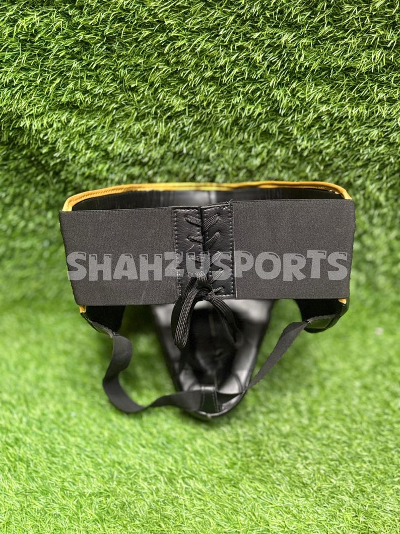 Puede incluir: Protector atl&eacute;tico negro con ribete dorado y el texto "SHAHZUSPORTS" en la cintura el&aacute;stica. El protector tiene un cierre de cordones y est&aacute; dise&ntilde;ado para la protecci&oacute;n deportiva. El fondo es verde.