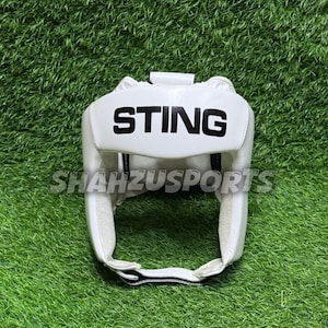 Op de afbeelding: Witte bokshoofdbescherming met het woord "STING" in zwarte blokletters over het voorhoofd. De hoofdbescherming heeft een kinband en oor bescherming. De tekst "SHAHZUSPORTS" is eronder gedrukt.