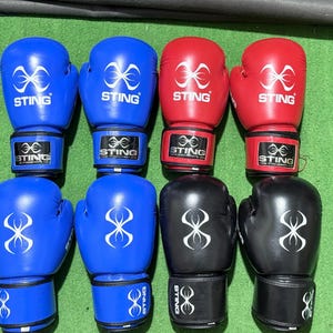 Op de afbeelding: Acht bokshandschoenen in blauw, rood en zwart. Elke handschoen heeft een wit logo en het woord "STING" erop. De handschoenen zijn gerangschikt in twee rijen van vier op een groene ondergrond.