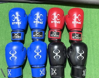Op de afbeelding: Acht bokshandschoenen in blauw, rood en zwart. Elke handschoen heeft een wit logo en het woord "STING" erop. De handschoenen zijn gerangschikt in twee rijen van vier op een groene ondergrond.