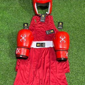 May include: A complete red boxing set, including a head guard with "STING" text, gloves, a tank top, and shorts. The gloves and headgear have a white logo. The set is displayed on a green surface.