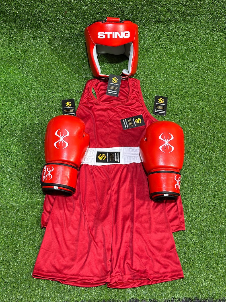May include: A complete red boxing set, including a head guard with "STING" text, gloves, a tank top, and shorts. The gloves and headgear have a white logo. The set is displayed on a green surface.